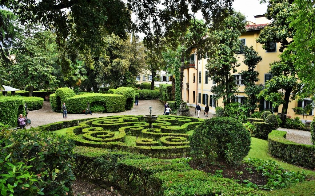 Giardino con siepe tagliata in modo geometrico, statue e casa di lato.