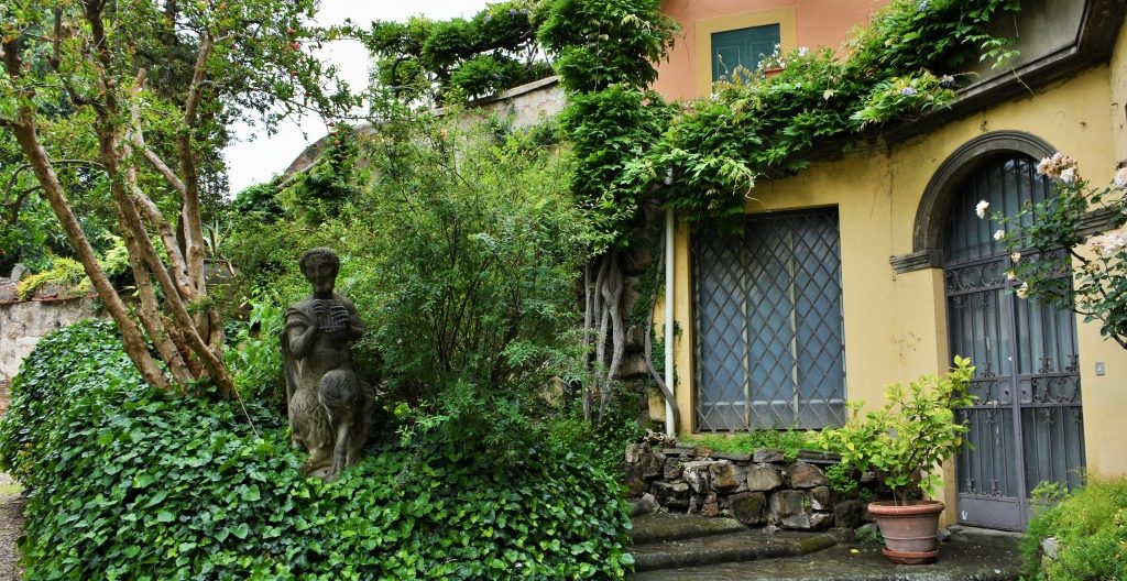 Statua di pietra raffigurante un musico tra l'edera e una scala, con un palazzo giallo e arancione.