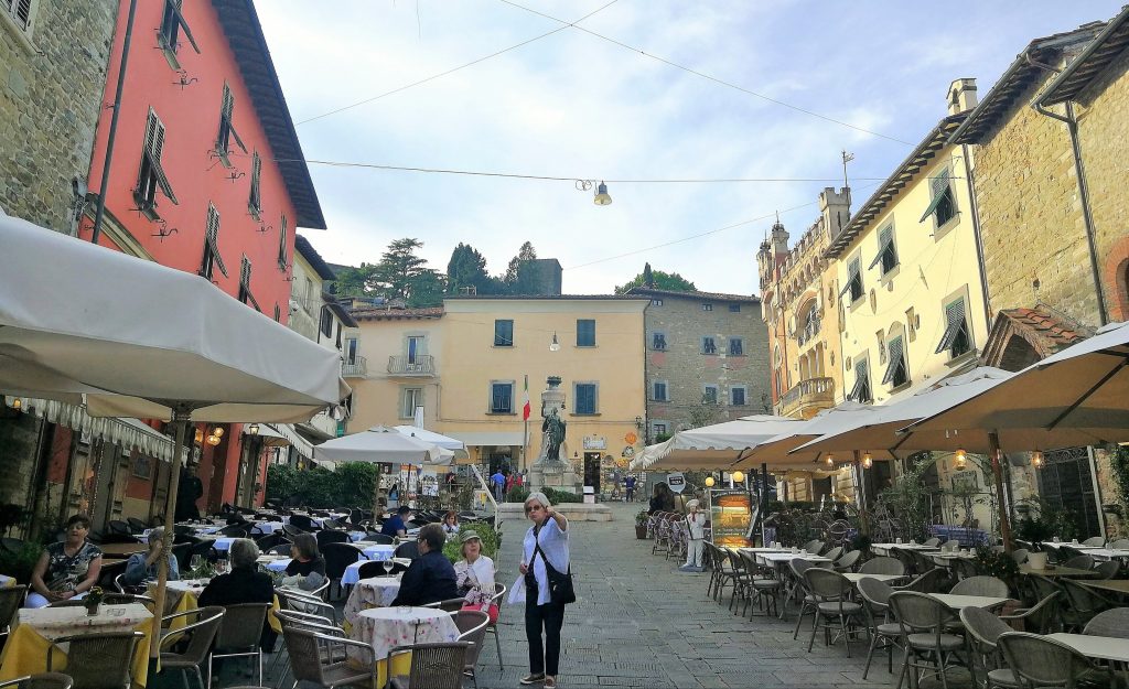 Piazza piena di tavoli e ombrelloni, alcune persone.