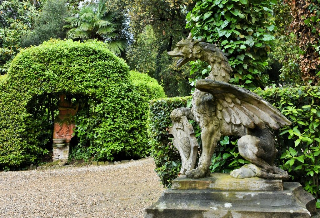 Statua di un drago di profilo con sfondo di siepi e alberi verdi e una statua di terracotta.