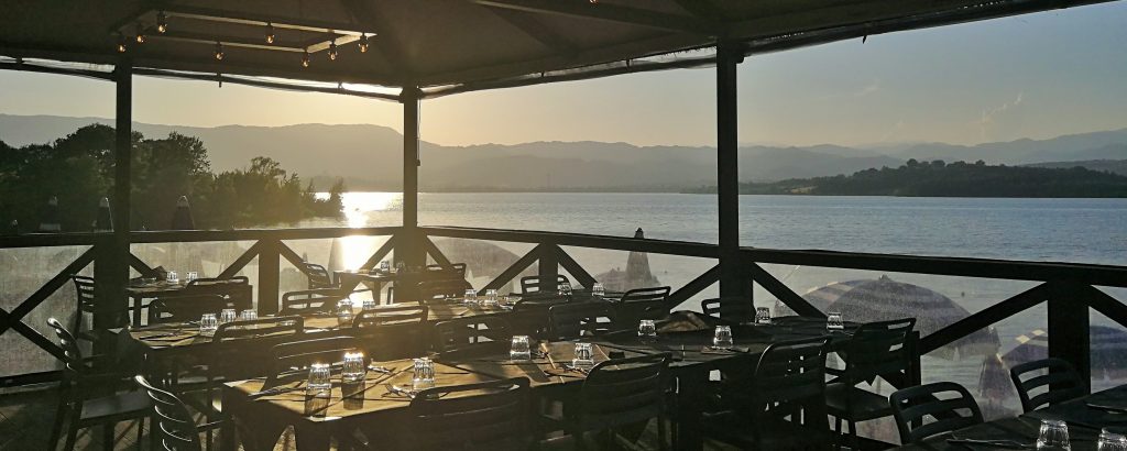 Terrazza con tavoli apparecchiati, sfondo del lago e vegetazione. Il sole sta tramontando e si specchia sull'acqua.