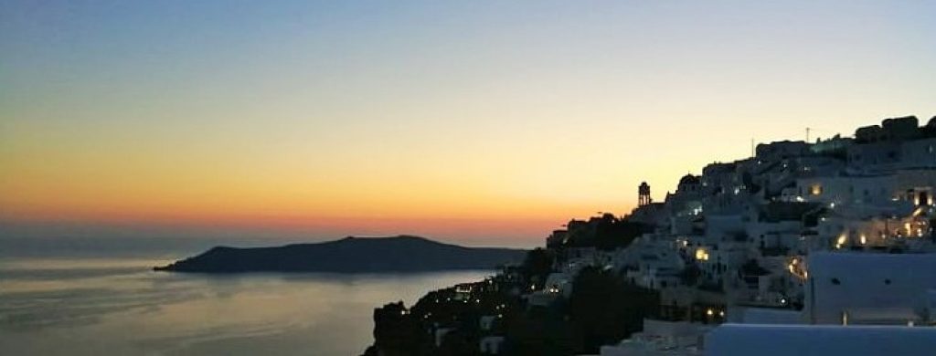 Tramonto sul mare; borgo arroccato di case bianche con alcune luci accese e sullo sfondo un'isola scura.