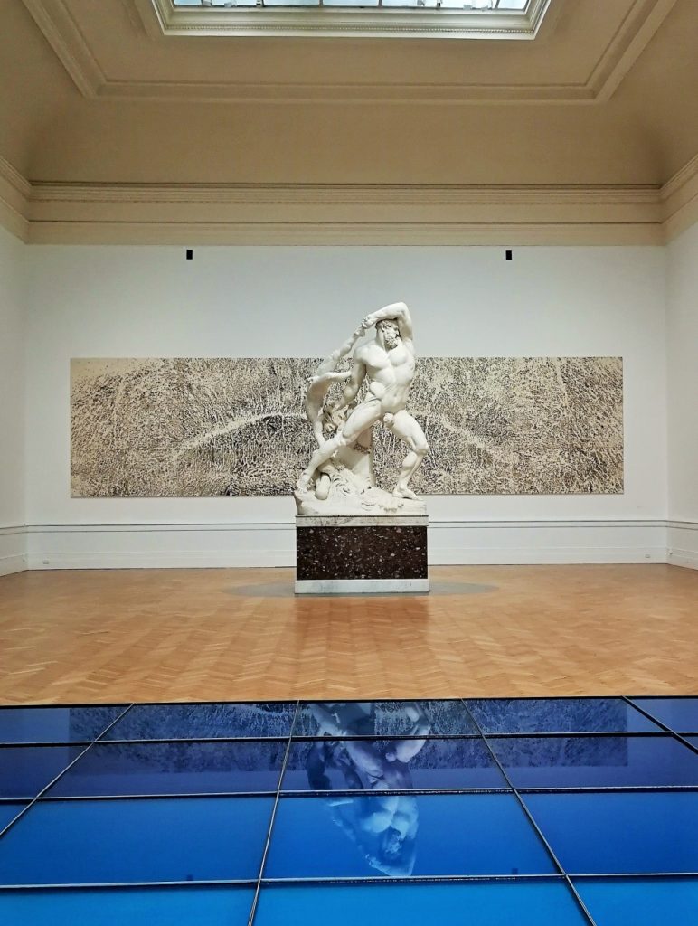 In primo piano un'opera d'arte raffigurante il mare, in diversi riquadri azzurri sul pavimento; su di essa si specchia la statua di Ercole del Canova. Dietro un'opera a parete che sembra sabbia.
