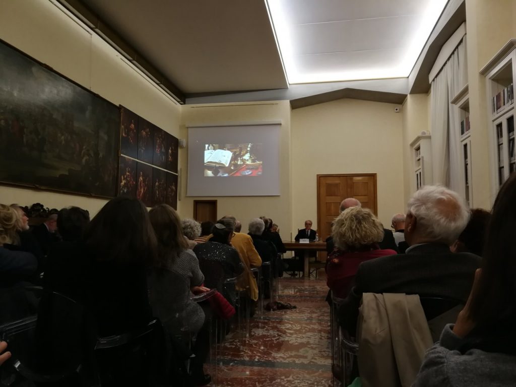 Sala colma di persone sedute, proiettore con un'immagine e tre relatori.