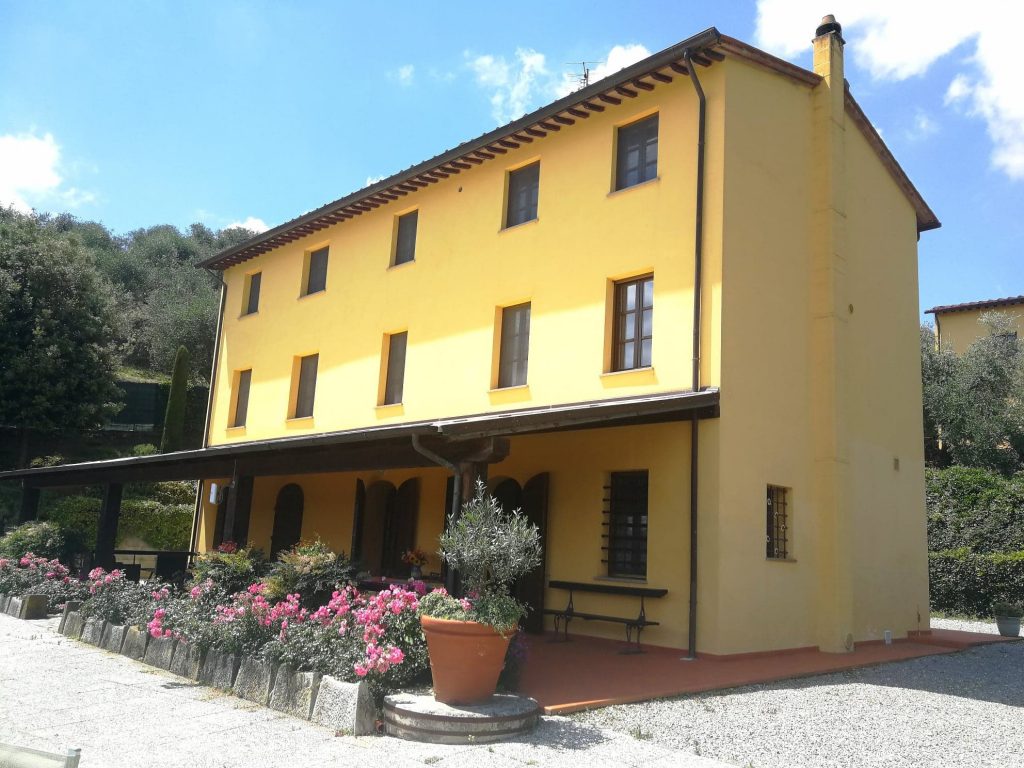 Casa gialla di grandi dimensioni, tettoia con tavolo e sedie, piante e fiori in vasi.