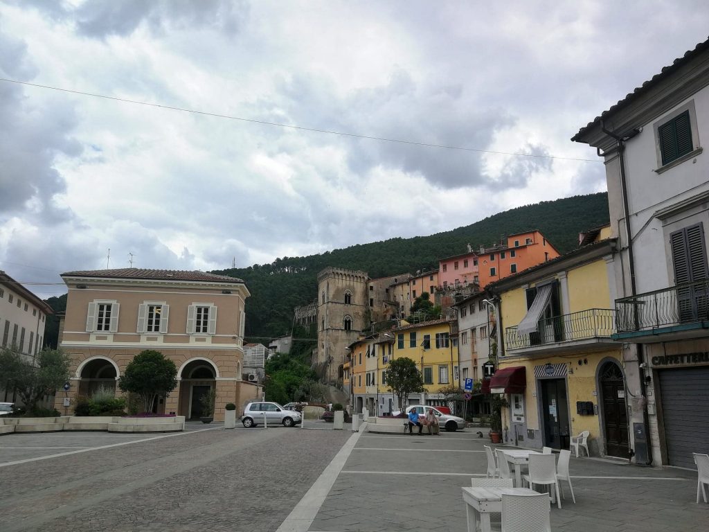 Piazza di Buti con tavolini bianchi, edifici colorati, il castello e la collina sullo sfondo.