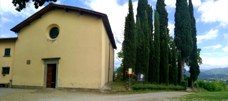 Chiesa intonacata, cipressi e scorcio di paesaggio collinare.
