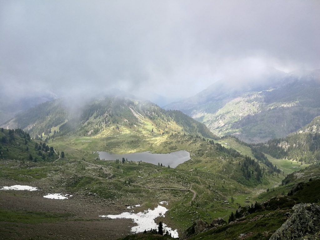 Montagne con lago, ghiaccio non sciolto, prati verdeggianti, nuvole basse.