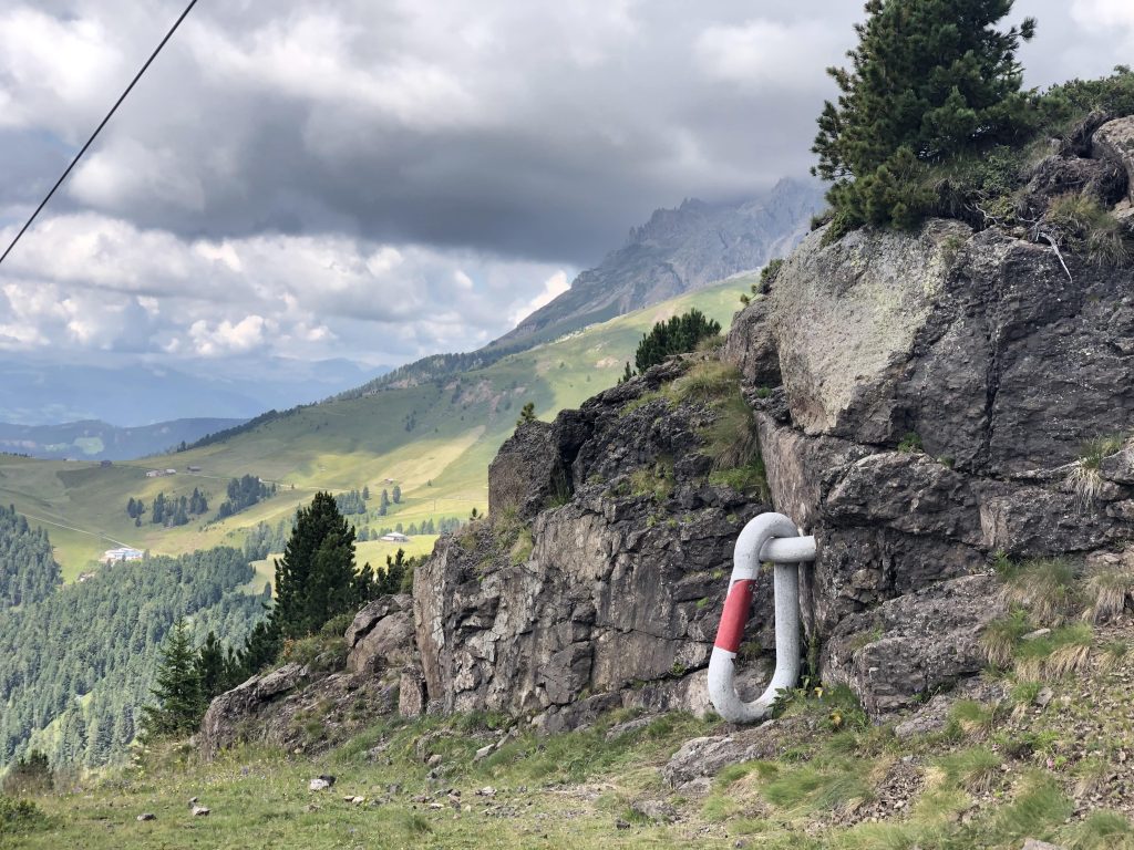 Panorama sulle dolomiti con montagne, boschi e prati verdeggianti; al centro un'opera d'arte, Sicurezza.