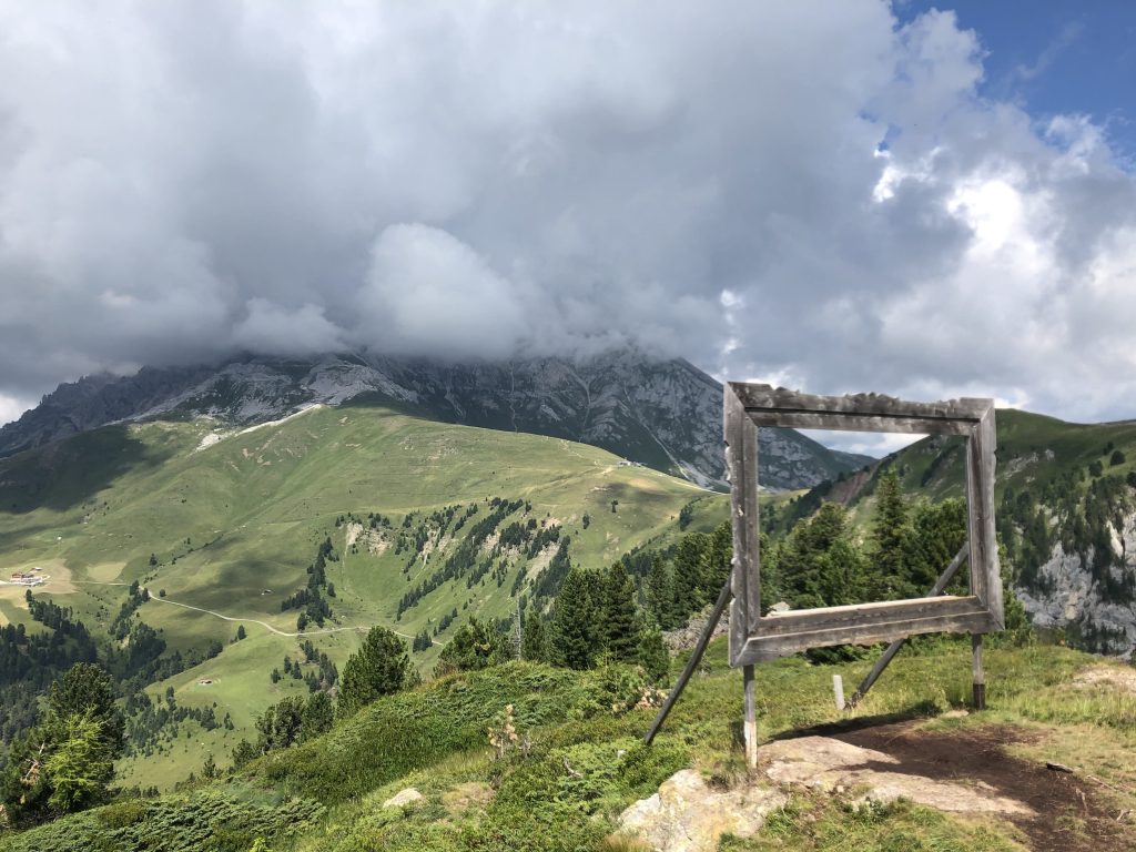Panorama sulle dolomiti con montagne, boschi e prati verdeggianti; al centro un'opera d'arte, Natura Viva.