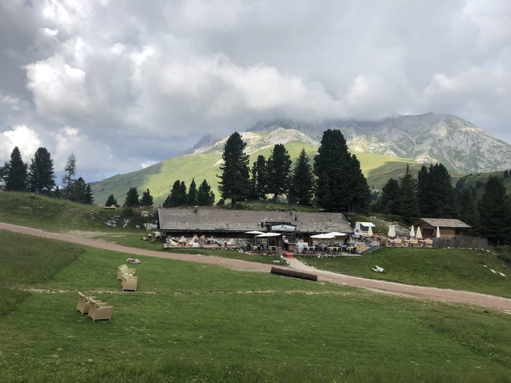 Panorama sulle dolomiti con montagne, boschi e prati verdeggianti; al centro uno chalet.
