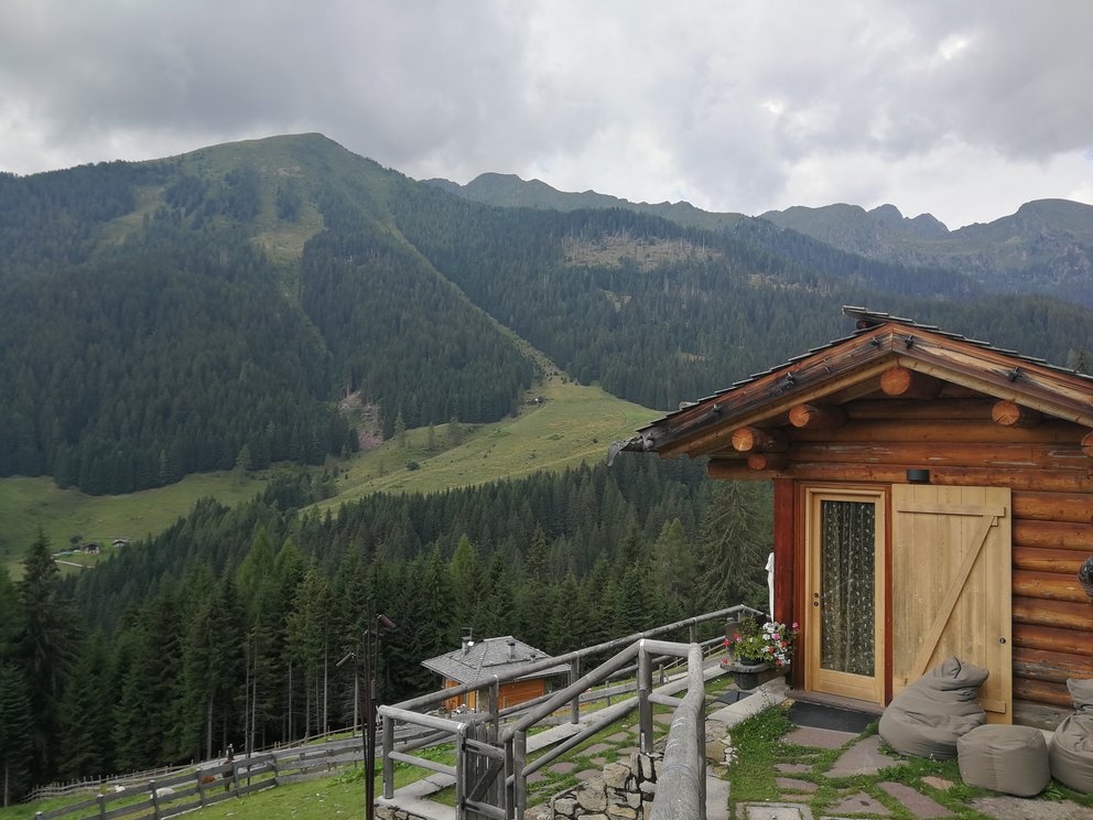 Piccola casa in legno, sullo sfondo boschi e montagne; cielo nuvoloso.