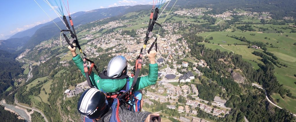 Parapendio biposto in volo sulla città di Cavalese, si vedono anche prati verdi e montagne.