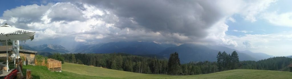 Panorama monti con nuvole, verde, prato, struttura in legno Malga Malghette con tavoli e ombrelloni.