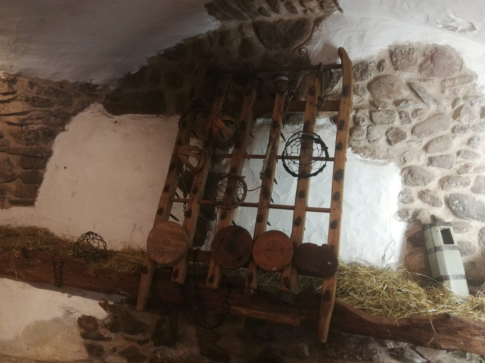 Museruole per vacche esposte su parete bianca e pietra, sotto mangiatoia con fieno.