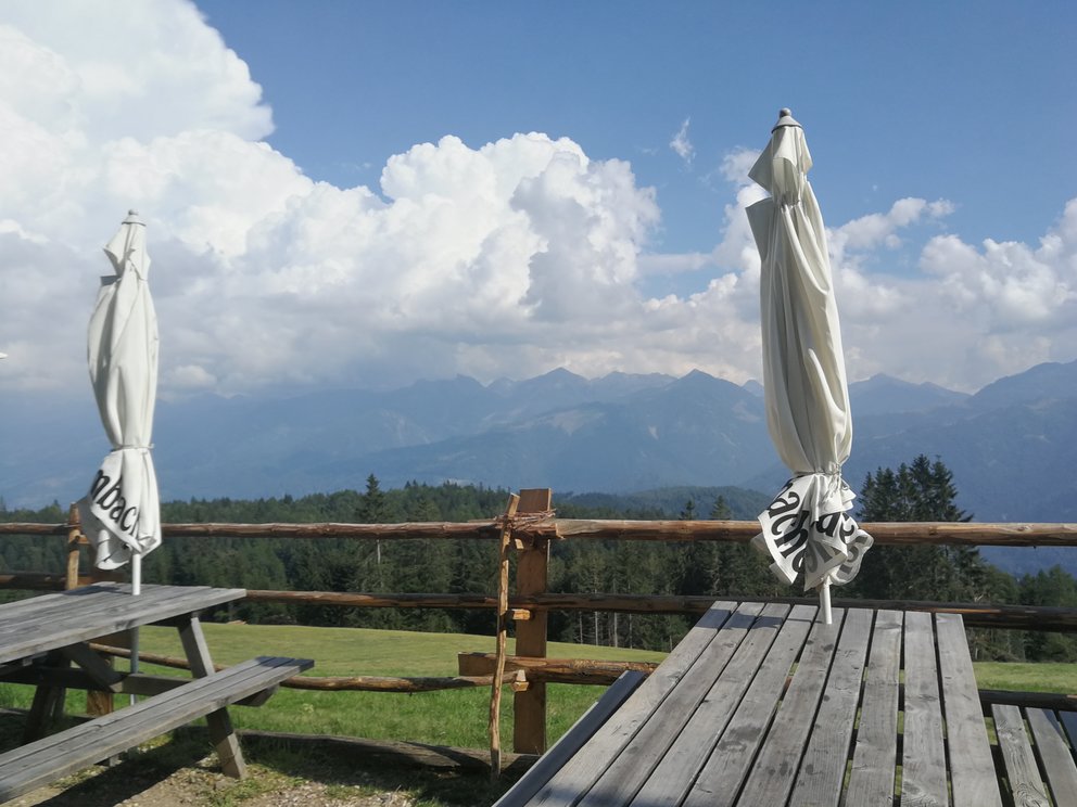 Monti con nuvole, verde, prato, struttura in legno Malga Malghette con tavoli e ombrelloni.
