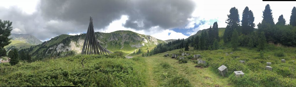 Panorama sulle dolomiti con montagne, boschi e prati verdeggianti; al centro un'opera d'arte, Teatro del Latemar.