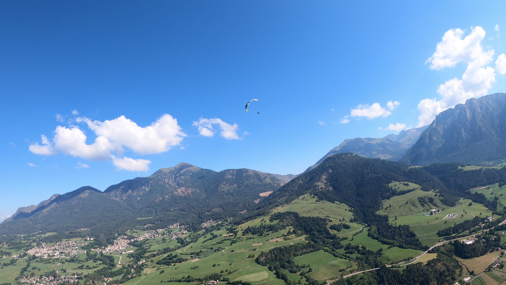 Panorama sulla valle con paesi e prati, montagne sullo sfondo. Contro il cielo, si vede un altro parapendio.