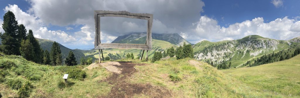 Panorama sulle dolomiti con montagne, boschi e prati verdeggianti; al centro un'opera d'arte, Natura viva.