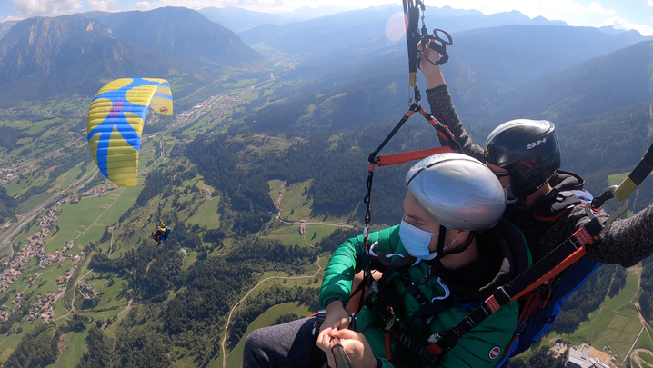 Parapendio biposto con Lorenzo e altro ragazzo, sotto si vede un altro parapendio e il panorama sulla valle: prati, case e montagne.