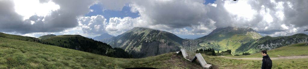 Panorama sulle dolomiti con montagne, boschi e prati verdeggianti; al centro un'opera d'arte.