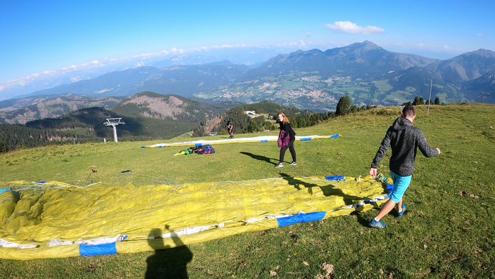 Vele dei parapendio distese con ragazzi che le preparano e Federica che saluta. Sullo sfondo il panorama sulla valle: boschi, monti e città.