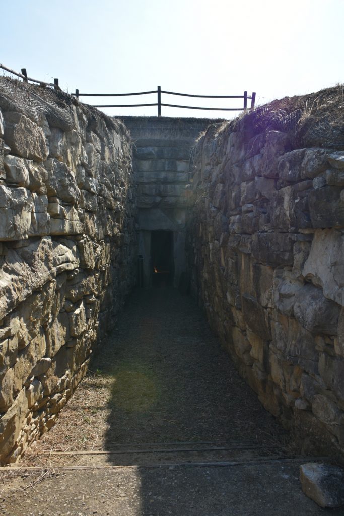 Corridoio di rocce e alla fine porta architravata, sopra staccionata e cielo.