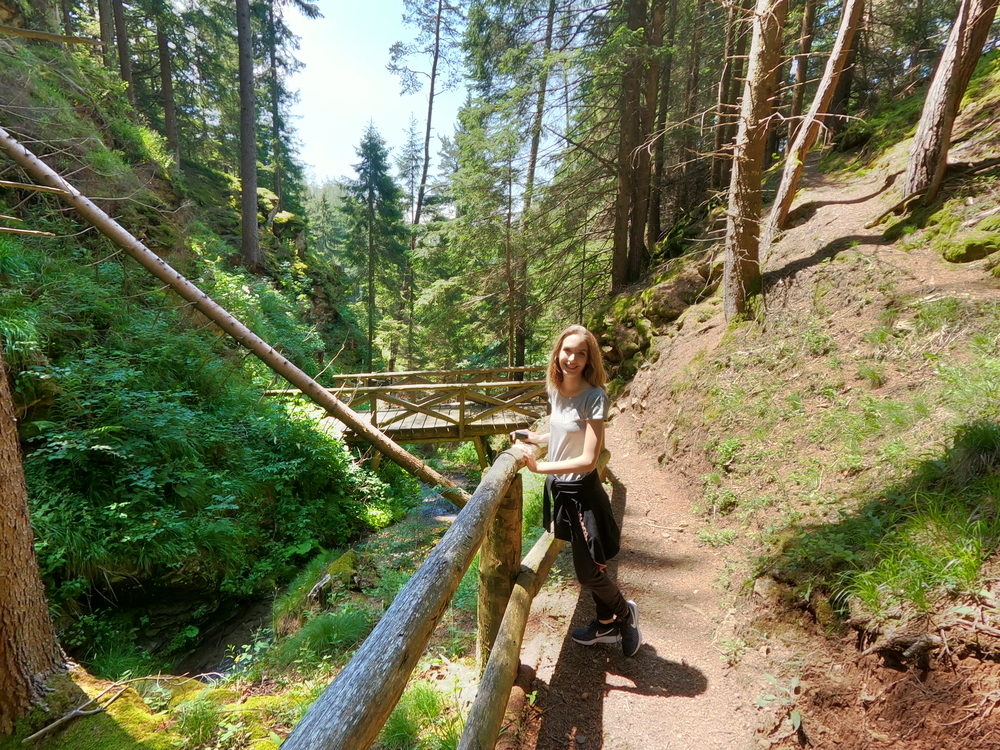 Percorso delle cascatelle - Federica appoggiata alla staccionata in legno, ponte in legno, alberi e in basso il corso d'acqua.