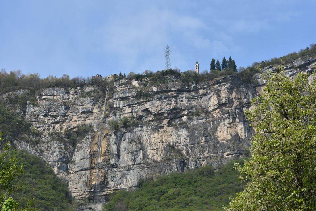 Cascata tra le rocce, alberi e in cima al dirupo spunta il campanile della chiesa Santi Filippo e Giacomo.