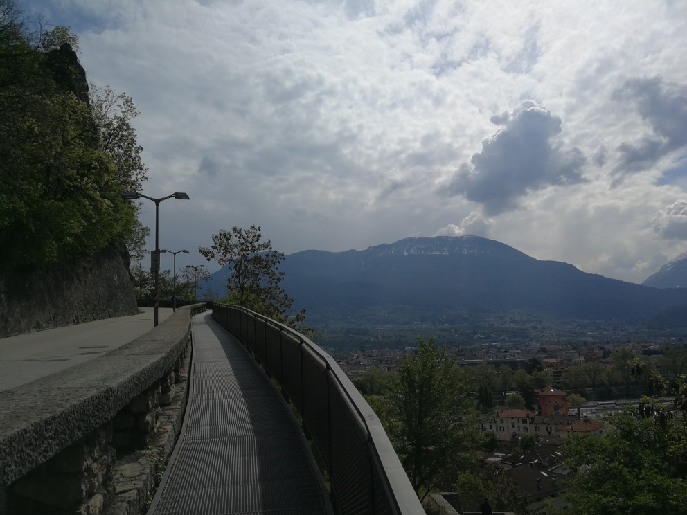 Percorso a piedi che costeggia la strada, bosco e montagne sullo sfondo.