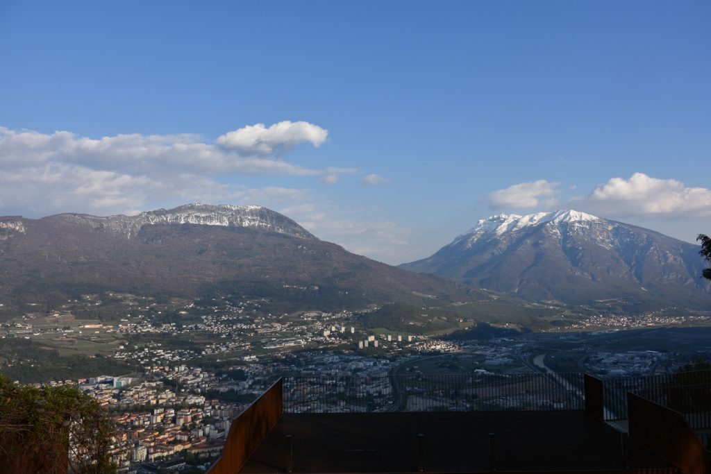 Panorama sulla città di Trento al tramonto, dall'alto, con montagne e fiume.