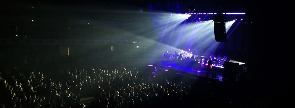Momento del concerto di Moroder: luci blu, orchestra, cantanti e Moroder sul palco, pubblico della platea parzialmente illuminato.