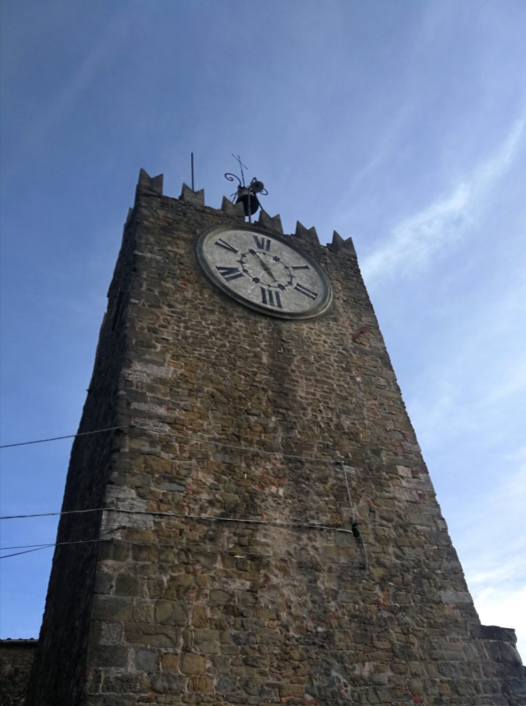 Torre medievale, orologio con fondo in marmo, merli e cielo azzurro.
