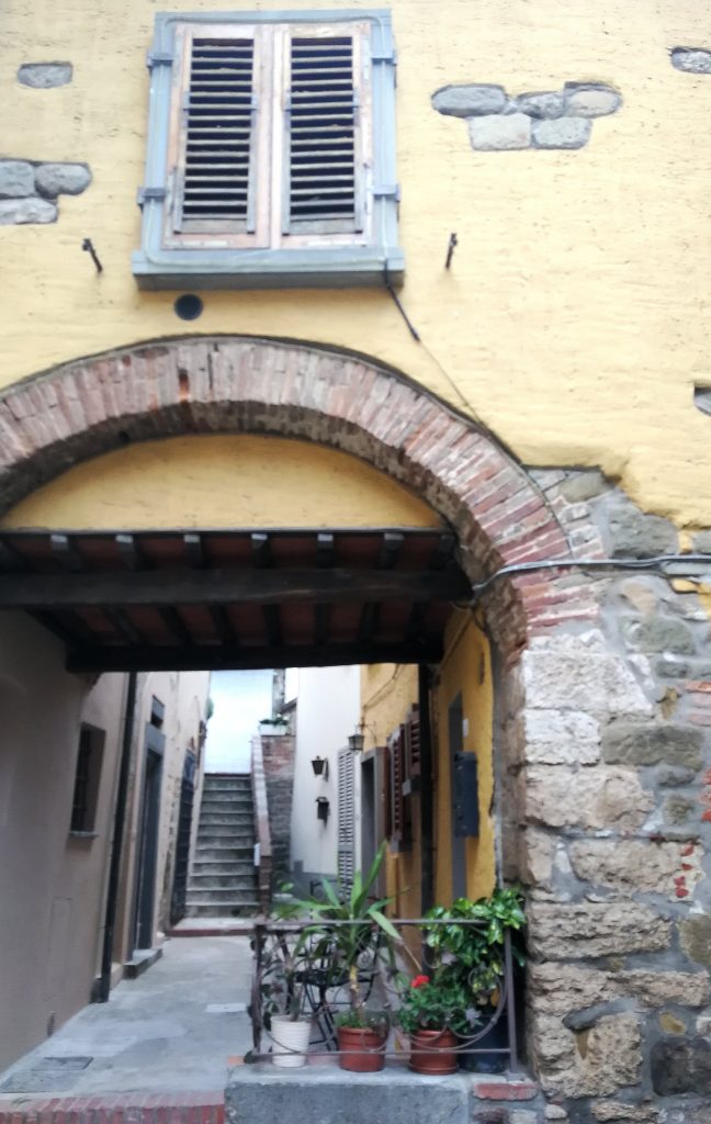 Casa in una via di Montecatini, arco a tutto sesto in mattoni rossi che apre su uno stretto viottolo, muro esterno della stanza giallo con finestre.