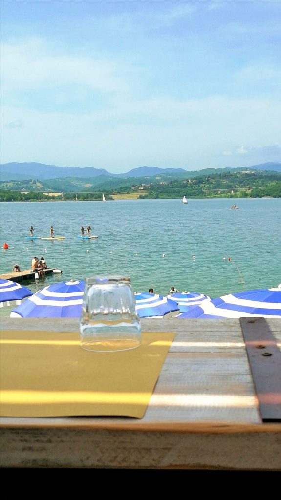 Tavolo in legno fatto a balcone apparecchiato con una tovaglietta e un bicchiere, con come sfondo il lago di Bilancino e le colline del Mugello.