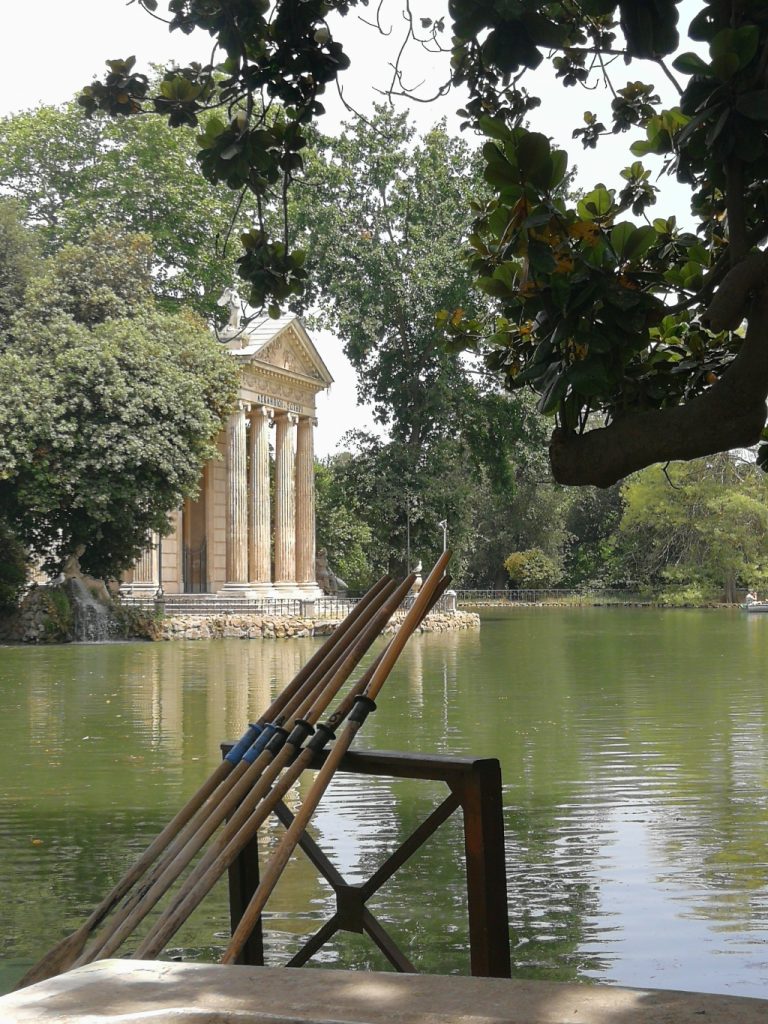 Tempio di architettura classica che sorge dalle acqua di un lago immerso nella vegetazione. In primo piano ci sono dei remi e una barca.
