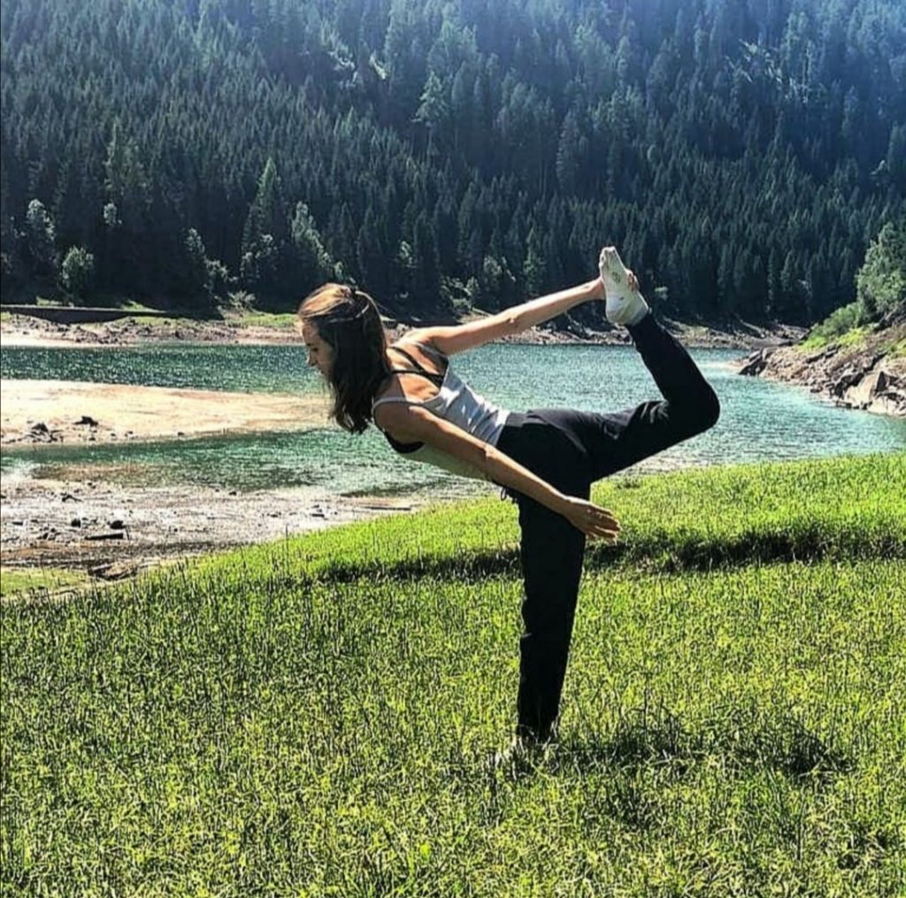 Ragazza che fa yoga tenendo il dorso del piede dex con la mano e restando in equilibrio. Sullo sfondo un lago e bosco.