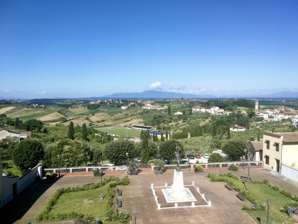 Panorama su piazza con statua e campagna verdeggiante.