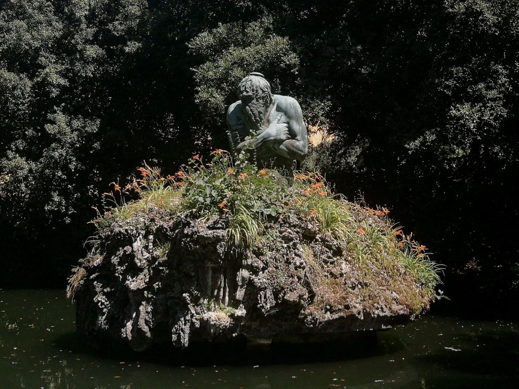 Statua in bronzo di un uomo che ha freddo, sopra un lago e dei fiori.