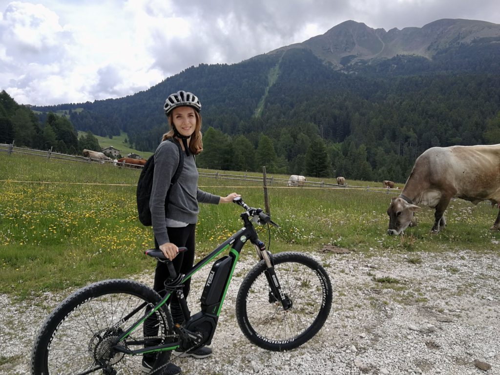 Federica con bici elettrica, mucche al pascolo e montagna con boschi.