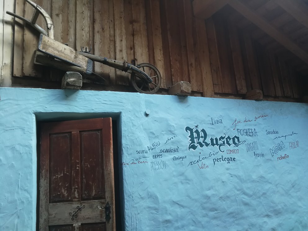 Entrata museo Carano: pareteazzurra con aratro in legno e scritte in dialetto.