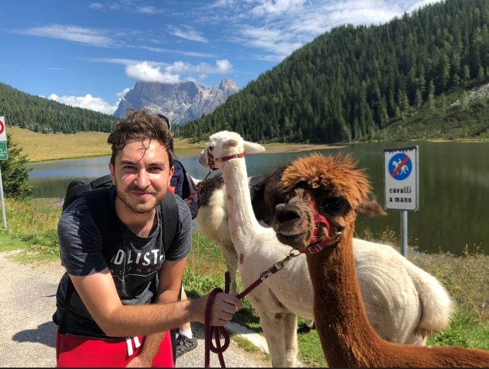 Lorenzo con due alpaca, uno bianco e uno marrone; sullo sfondo altre persone e lama, boschi e lago.