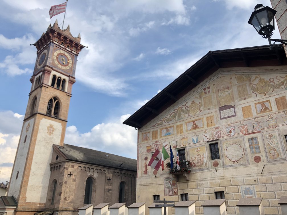 Palazzo della Magnifica Comunità di Fiemme, rinascimentale con affreschi sulla facciata, bandiere. Campanile di una chiesa.