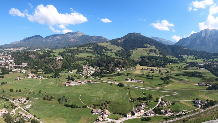 Panorama sulla valle visto dall'alto: paesi, prati verdi e boschi, montagne sullo sfondo.
