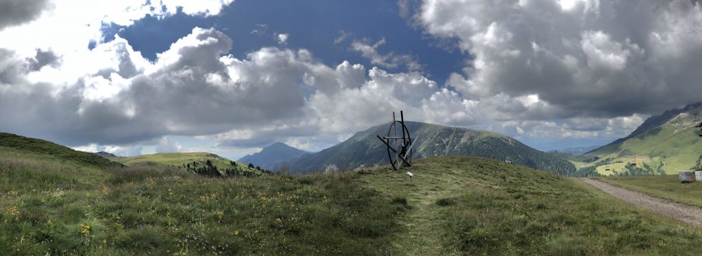 Panorama sulle dolomiti con montagne, boschi e prati verdeggianti; al centro un'opera d'arte, Il nodo della strega.