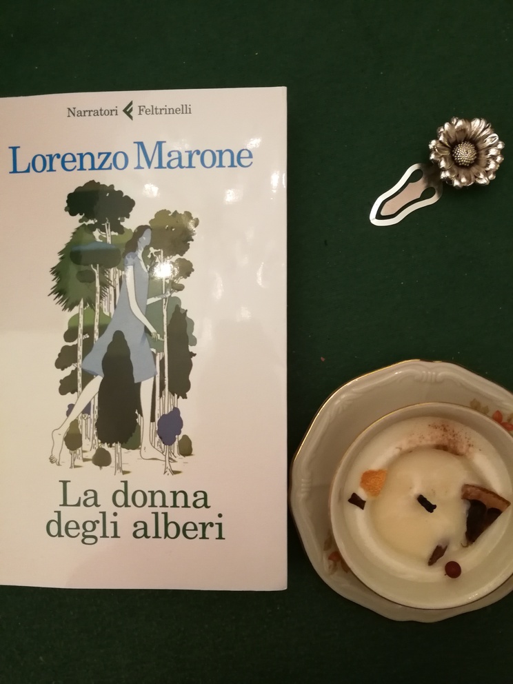 Copertina del libro La donna degli alberi con accanto una candela un po' consumata ricavata in una tazza da caffè con piattino e un fermalibro argento con un fiore.