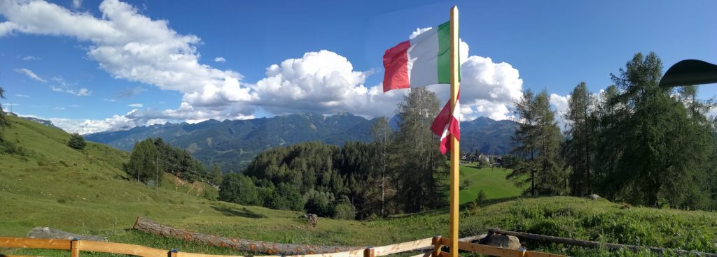 Chiesetta degli Alpini - staccionata, bandiere dell'Italia al vento, prati e panorama di montagne con alberi.